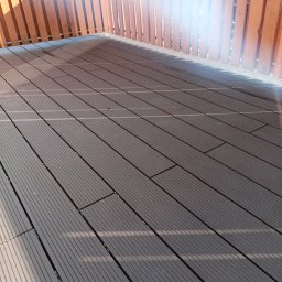 Dam-Bud Damian Przybylak - Nowoczesna zabudowa balkonu w Koninie z brązową deską kompozytową i drewnianą balustradą w ciepłym odcieniu, oświetlona promieniami słońca.