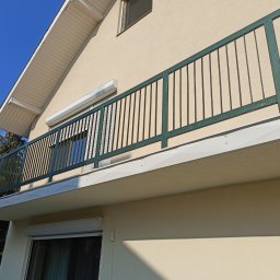 Dam-Bud Damian Przybylak - Zabudowany balkon z zieloną metalową balustradą, kremowa elewacja budynku i częściowo opuszczona roleta okienna.