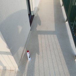 Dam-Bud Damian Przybylak - Biały balkon z szarą, nakrapianą posadzką, częściowo zacieniony przez balustradę, z butelką płynu do czyszczenia w rogu.