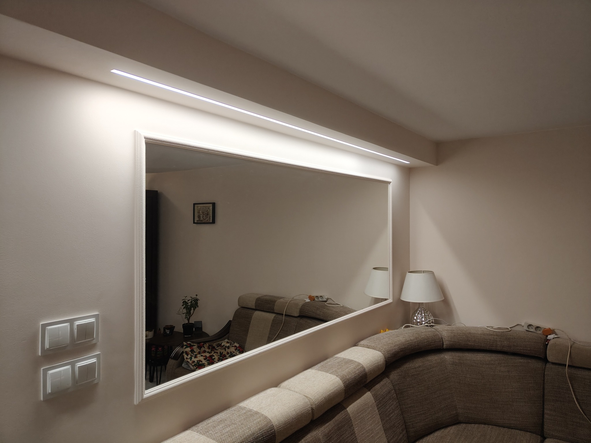 Duże lustro w białej ramie zamontowane na ścianie, odbijające część pokoju z kanapą i lampą, oświetlone ledowym paskiem nad lustrem, obok widoczne białe włączniki światła.