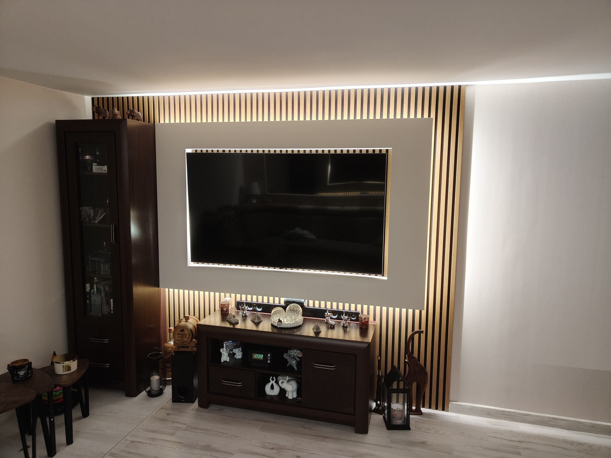 Salon z drewnianą ścianką działową za telewizorem, podświetloną listwą LED, ciemnymi meblami i dekoracjami.