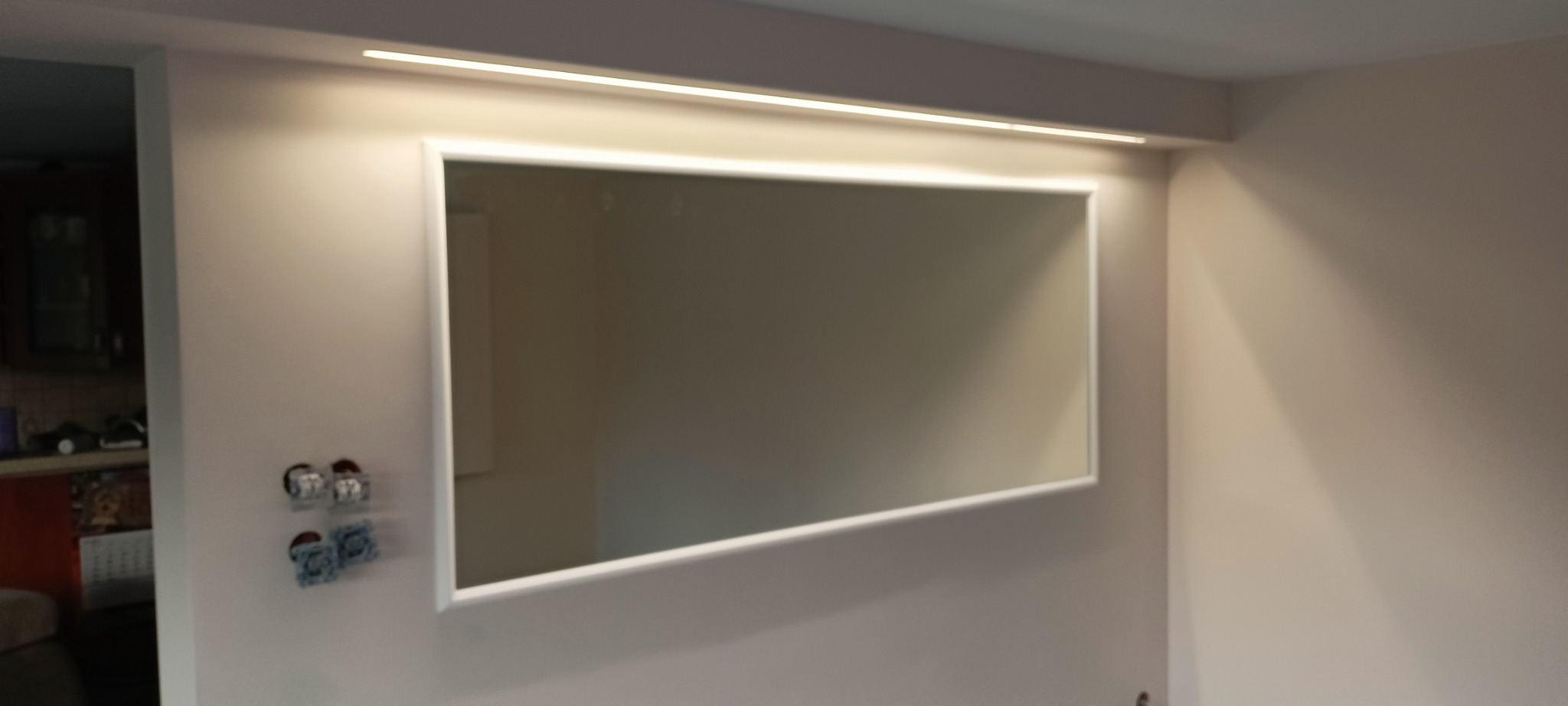 Wbudowane lustro w ramie z oświetleniem LED nad nim, widoczne gniazdka elektryczne po lewej stronie.