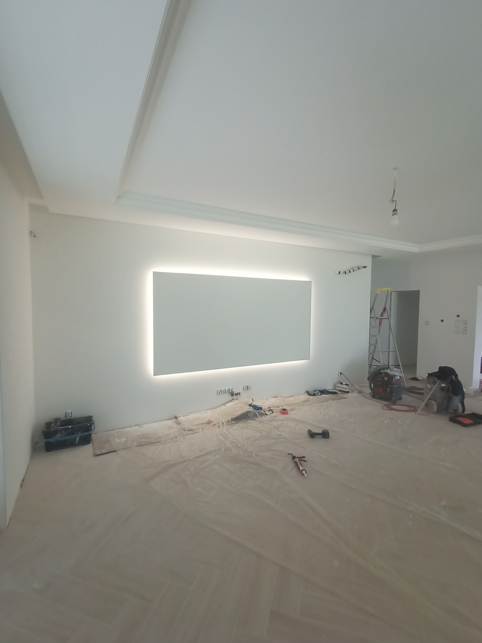 Wnętrze w trakcie prac wykończeniowych: białe ściany z podświetlanym prostokątnym panelem, zabezpieczona folią podłoga, drabina, narzędzia i sprzęt budowlany w przestronnym pomieszczeniu z listwami...