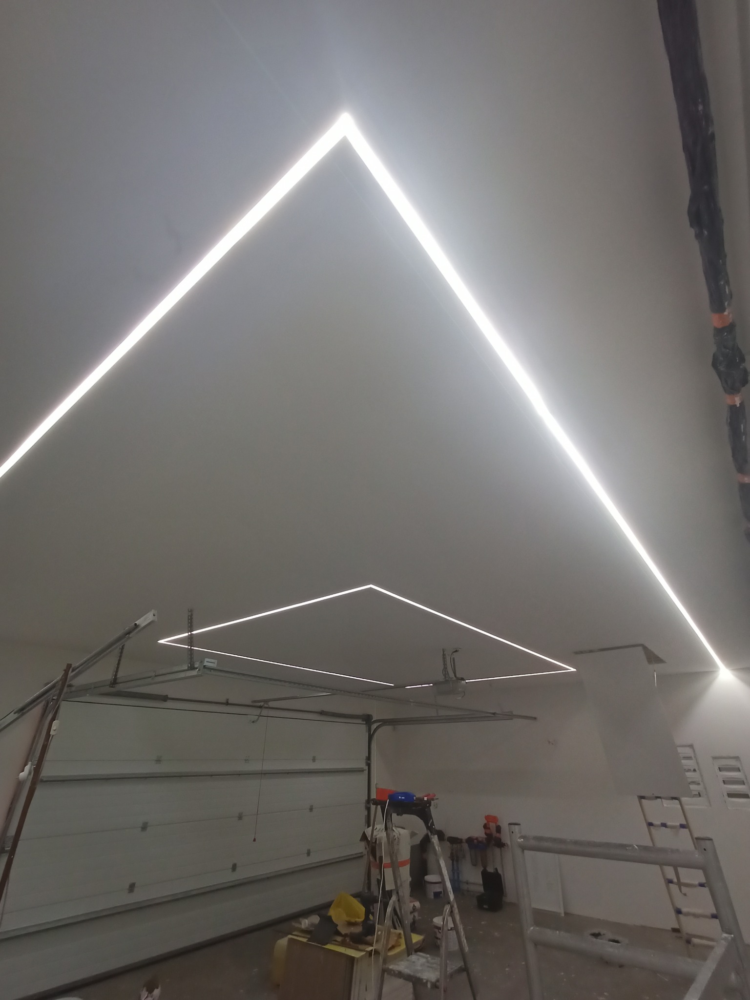 Nowoczesne oświetlenie LED w kształcie geometrycznym na suficie w garażu, widoczne drabiny i brama garażowa, surowy stan wykończenia.