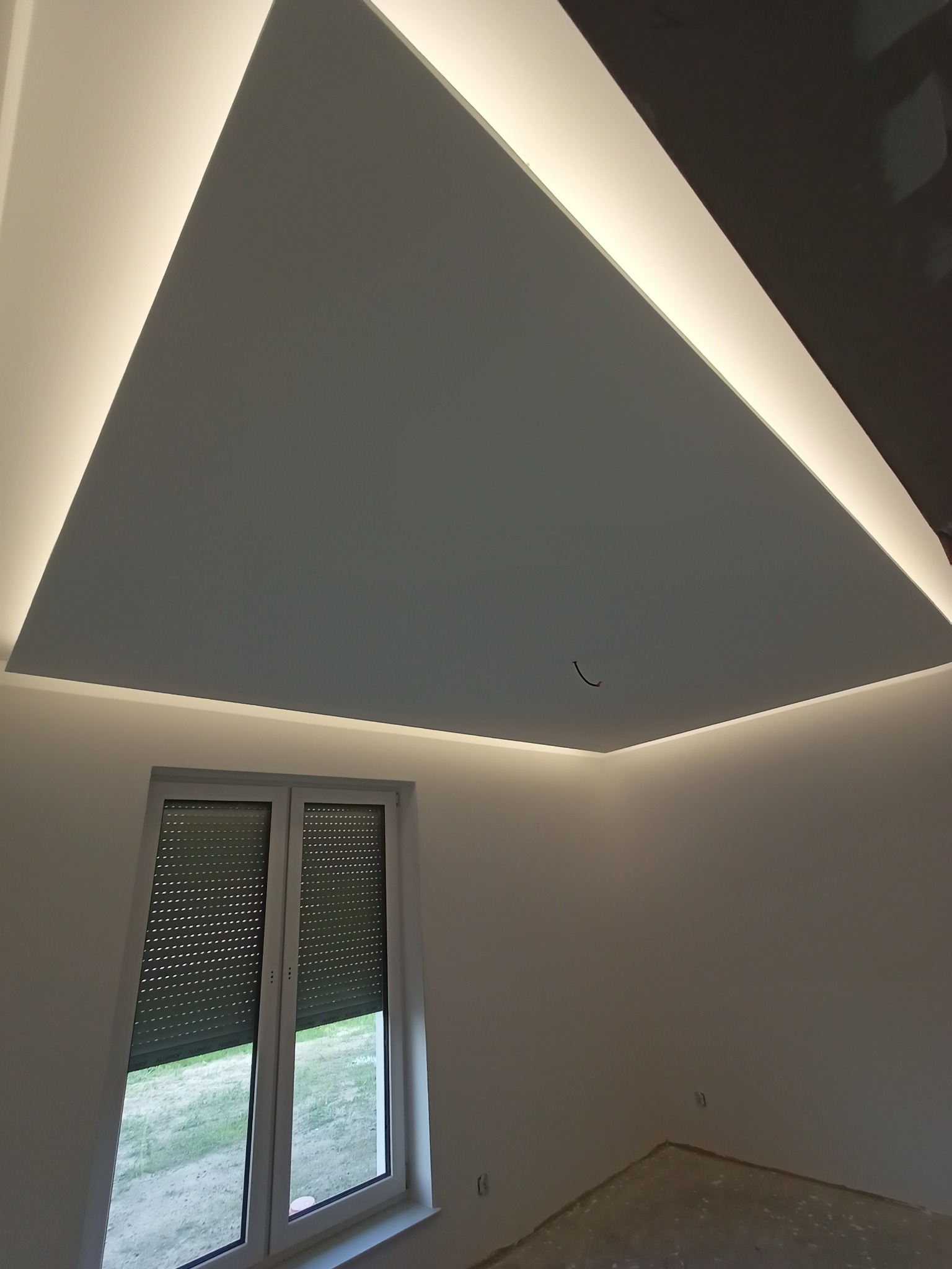 Pokój z podwieszonym sufitem z oświetleniem LED, widoczne okno z żaluzjami i fragment podłogi przed wykończeniem.