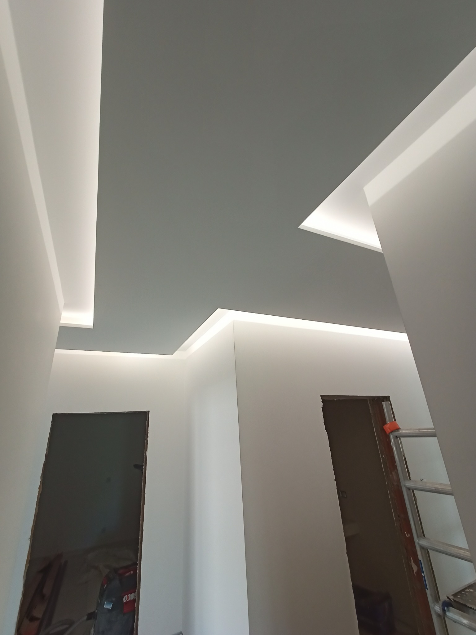 Nowoczesne wnętrze w trakcie prac wykończeniowych: szara, podwieszana część sufitu z oświetleniem LED, białe ściany, otwór drzwiowy bez drzwi i drabina w rogu pomieszczenia.