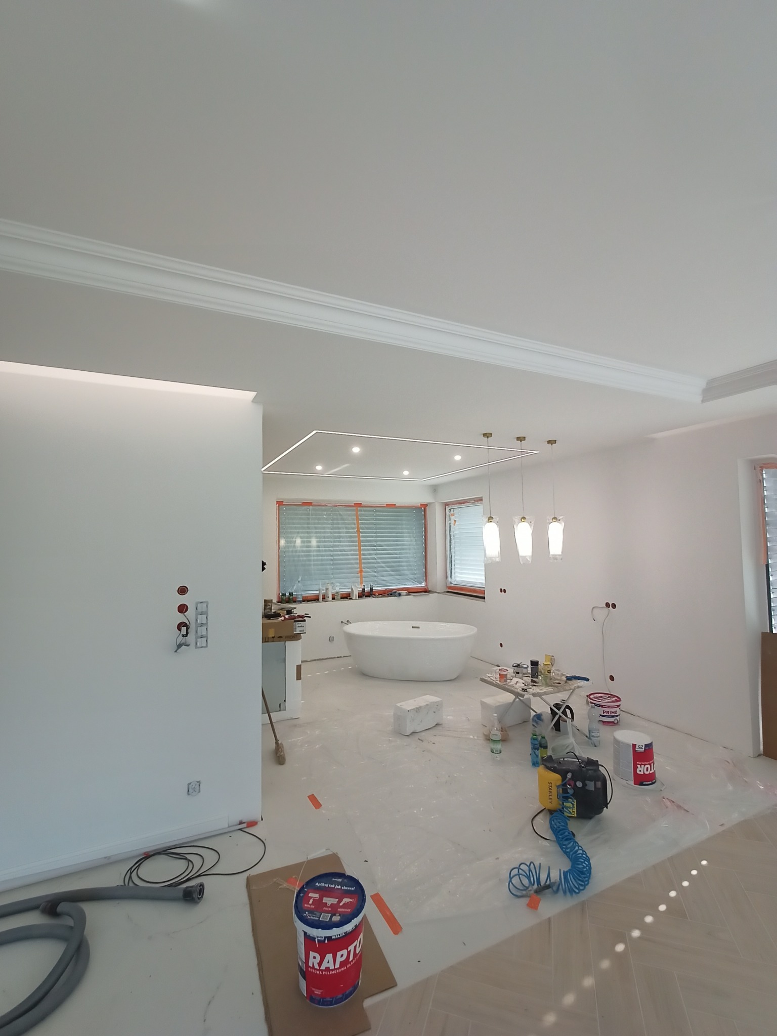 Wnętrze łazienki w trakcie prac wykończeniowych: widoczna wanna wolnostojąca, puszki z farbą, kompresor, osłonięta podłoga, listwy dekoracyjne pod sufitem i oświetlenie punktowe.