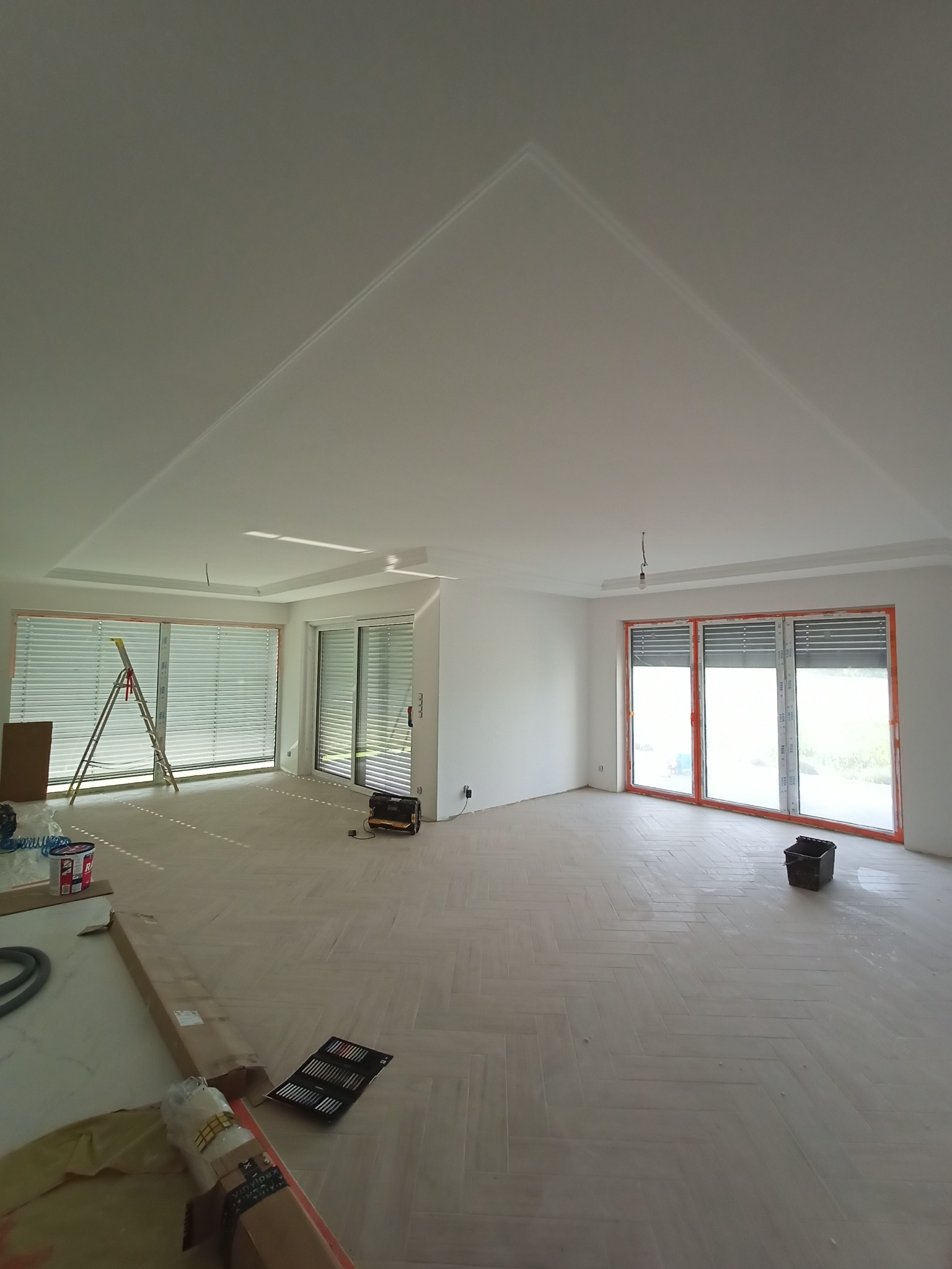 Jasne, przestronne wnętrze z ułożonym parkietem w jodełkę, świeżo pomalowanymi ścianami i sufitem z dekoracyjną listwą, widoczne okna z żaluzjami oraz drabina i narzędzia malarskie.