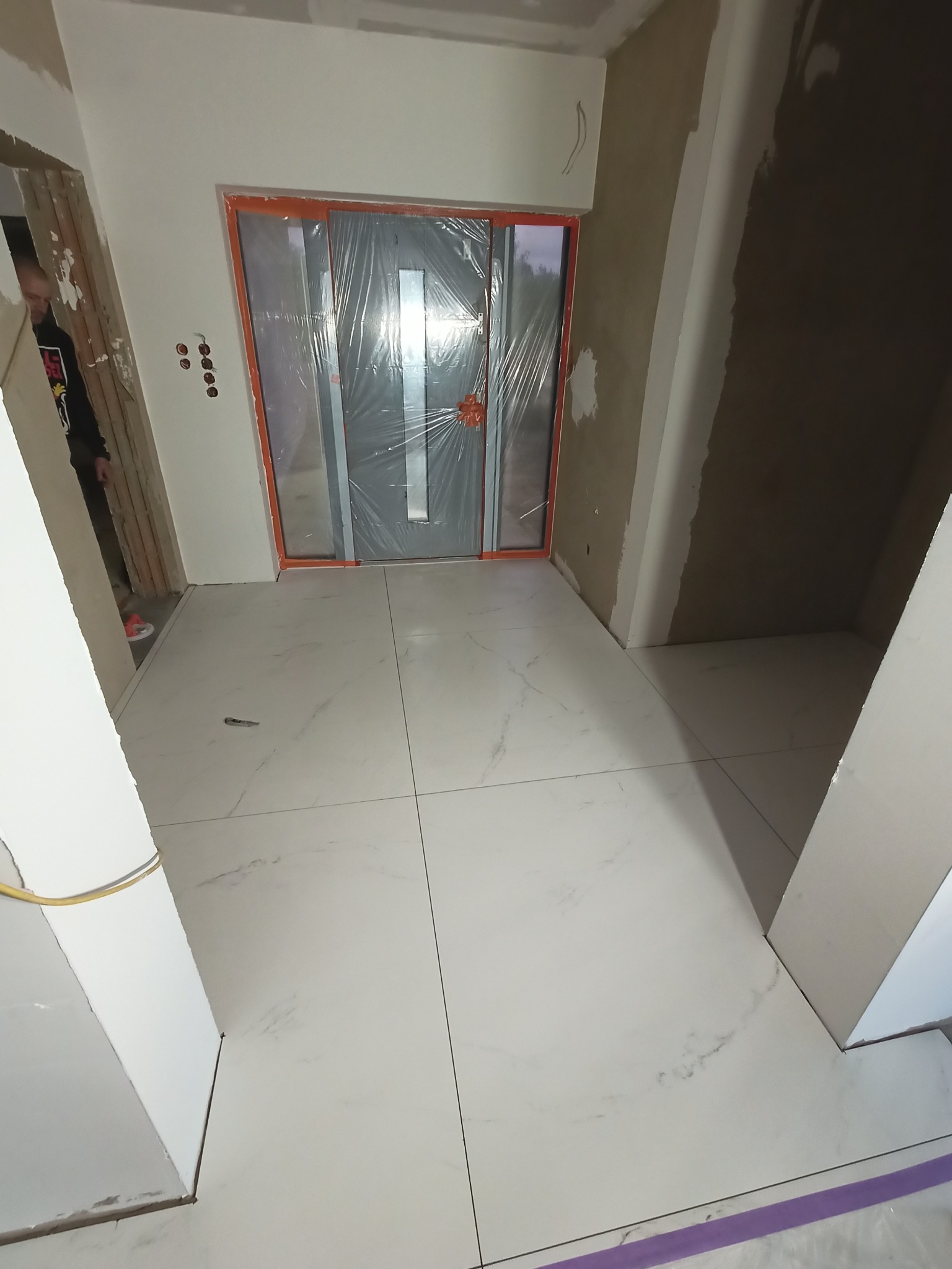 Nowe, białe płytki podłogowe imitujące marmur, ułożone w korytarzu podczas prac wykończeniowych, z zabezpieczonymi folią drzwiami wejściowymi i widocznymi śladami gładzi na ścianach.