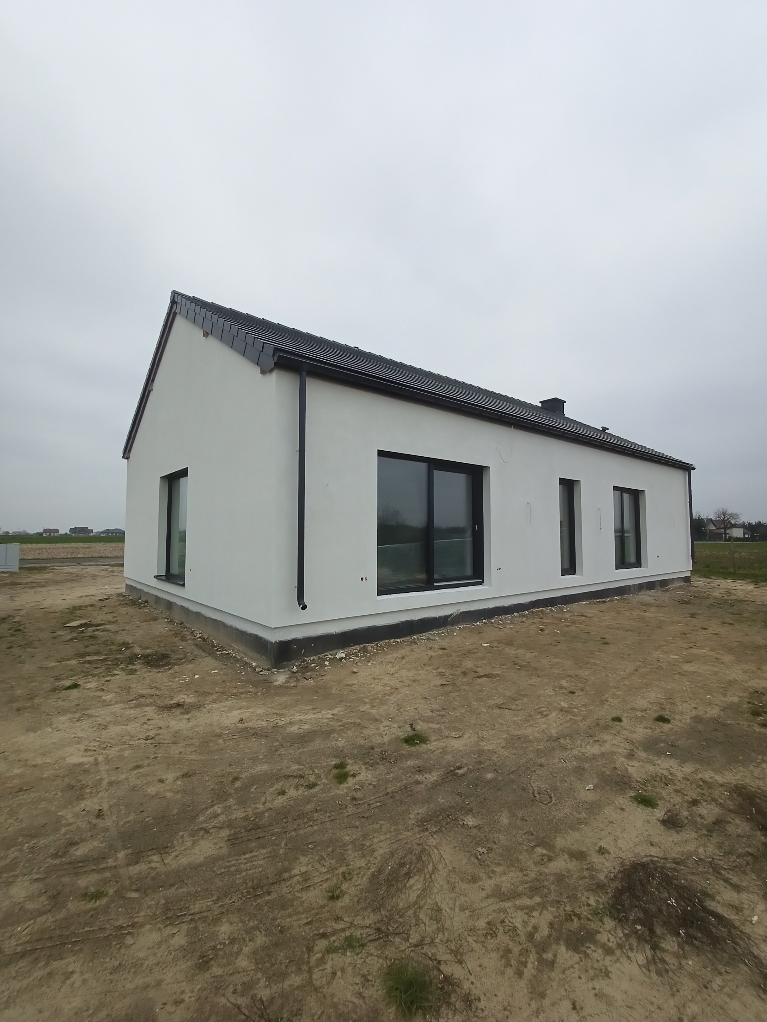 Nowoczesny, parterowy dom jednorodzinny z białymi ścianami zewnętrznymi, czarną dachówką i czarnymi ramami okiennymi, widoczny z zewnątrz na etapie wykończenia terenu.