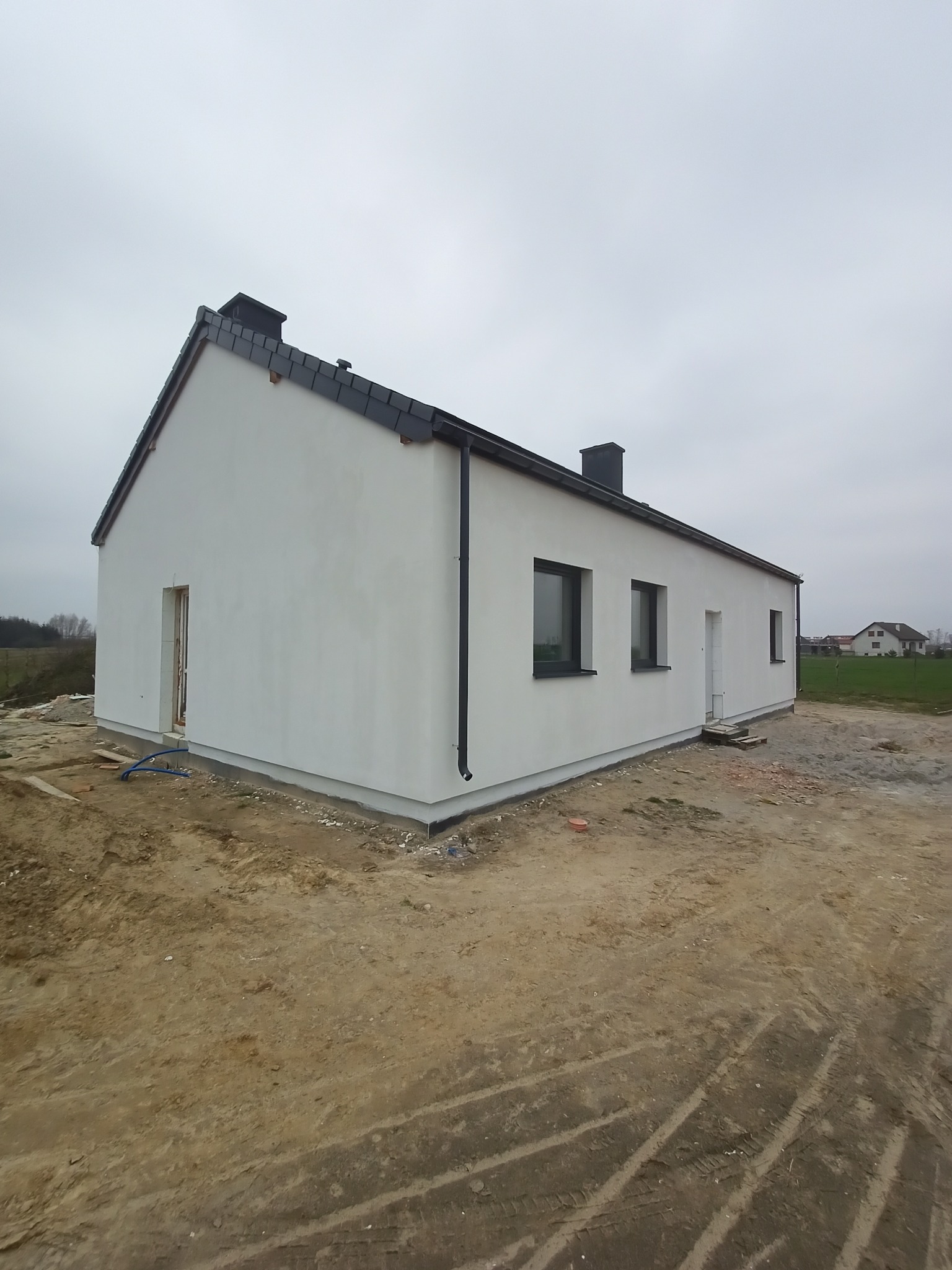 Jednopiętrowy dom z białymi ścianami i ciemnymi oknami, widziany z zewnątrz na działce w trakcie prac wykończeniowych, z widocznymi śladami opon na ziemi.
