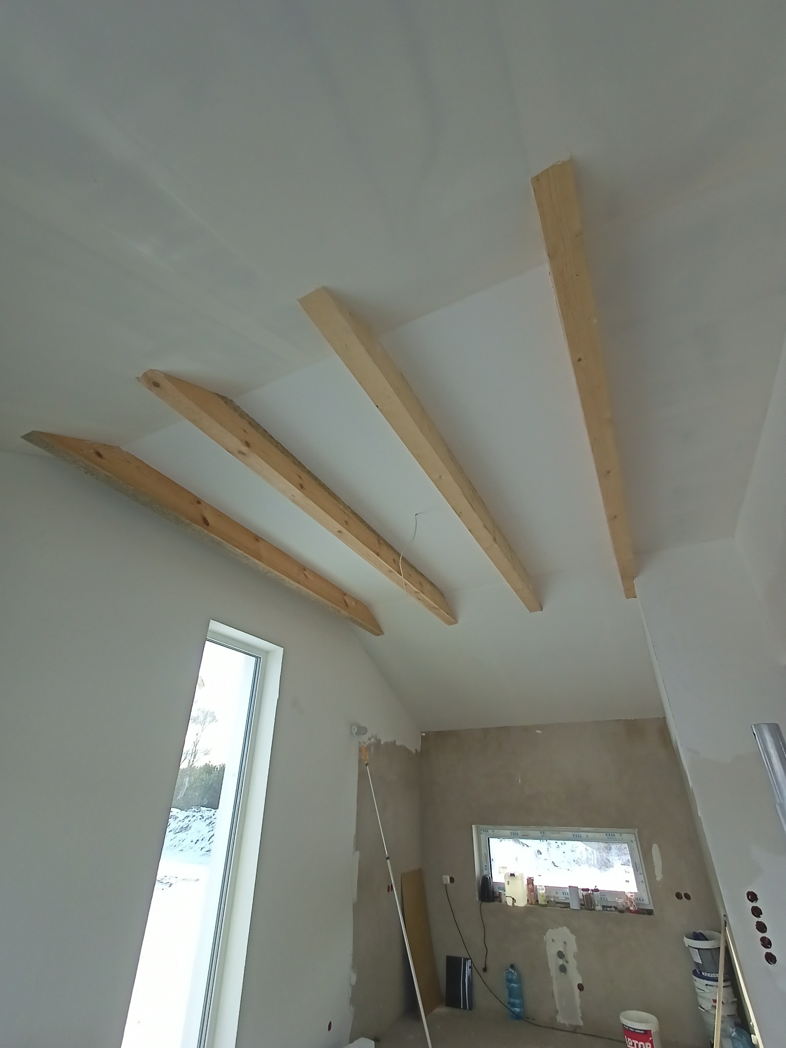 Wnętrze w trakcie prac wykończeniowych: surowy sufit z drewnianymi belkami, częściowo pomalowane ściany, widoczne okno i narzędzia malarskie.