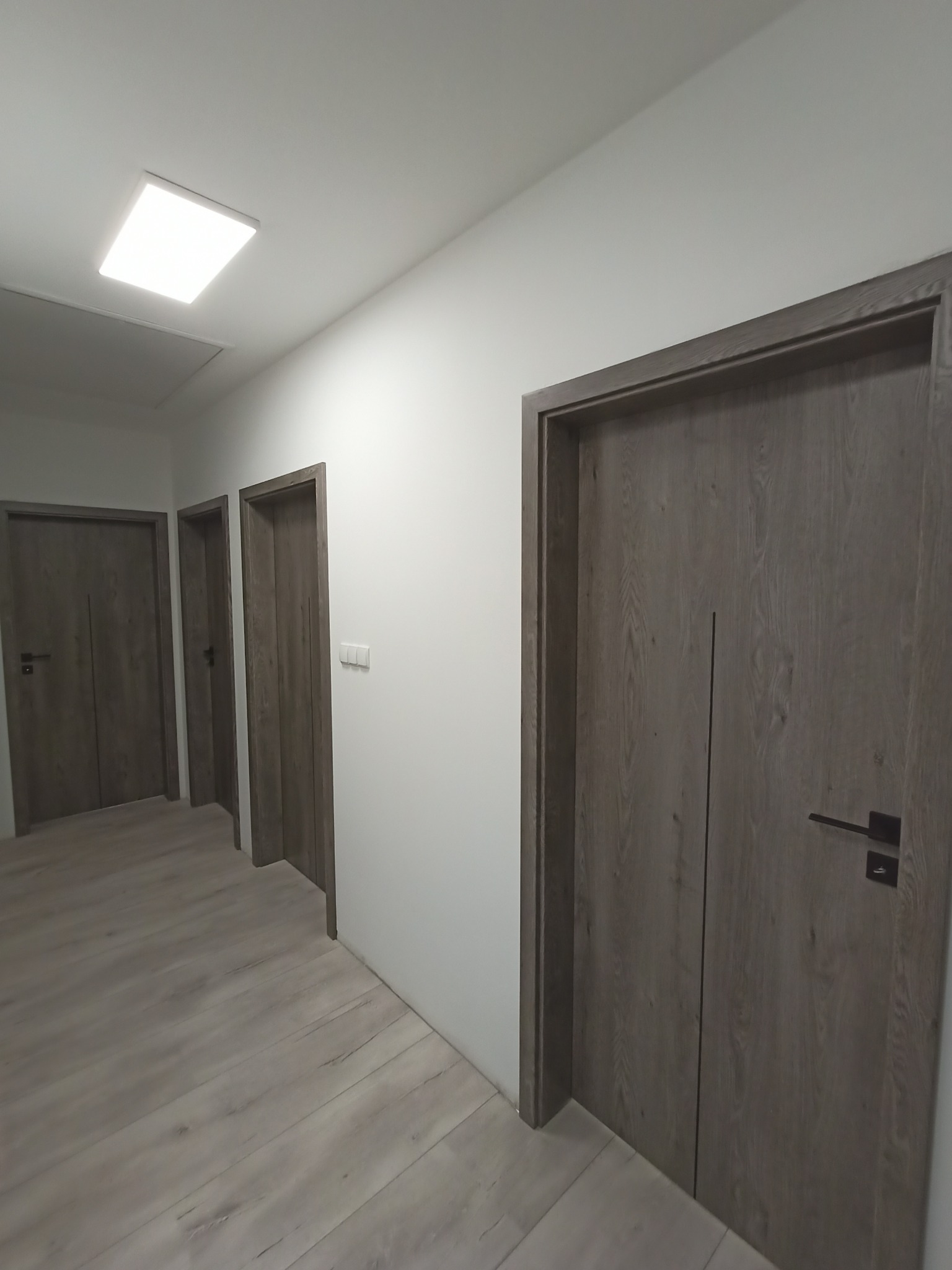 Nowoczesne, szare drzwi wewnętrzne z czarną klamką w minimalistycznym wnętrzu z panelami podłogowymi imitującymi drewno.