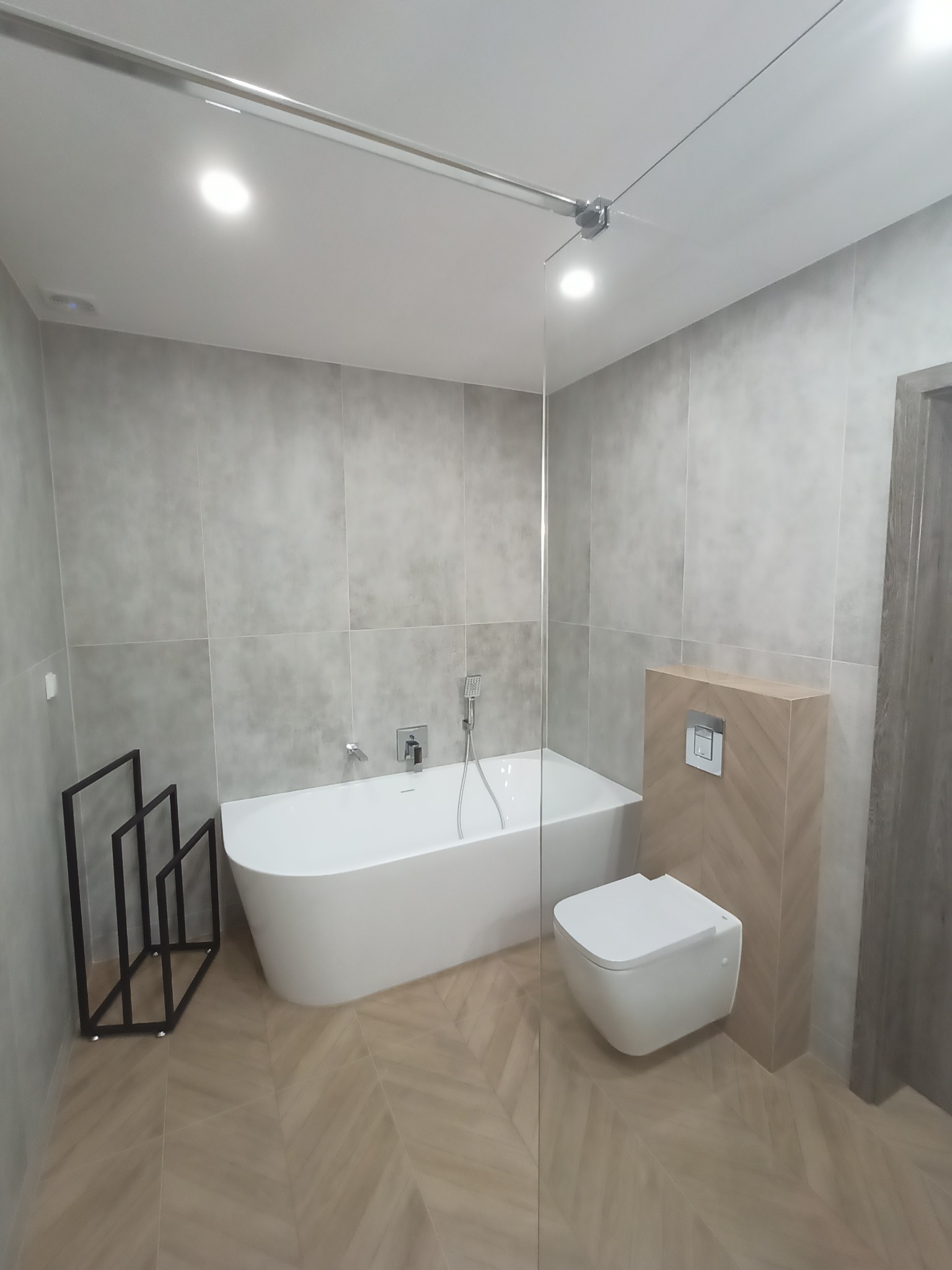 Nowoczesna łazienka z wolnostojącą białą wanną, podwieszaną toaletą i szklaną ścianką prysznicową, wykończona szarymi płytkami i podłogą w jodełkę.