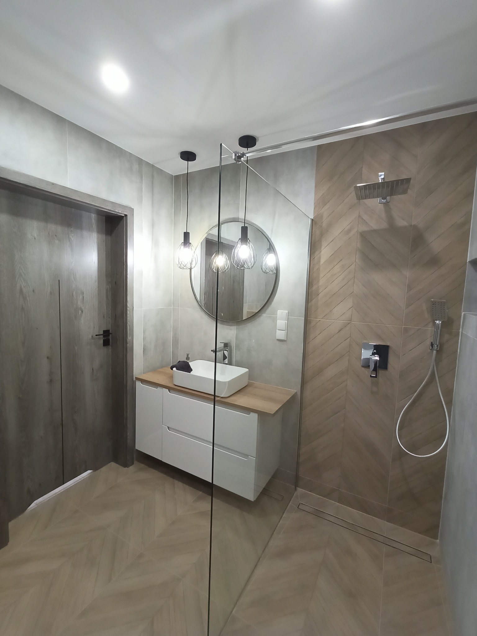 Nowoczesna łazienka z kabiną prysznicową typu walk-in, umywalką nablatową na białej szafce z drewnianym blatem, okrągłym lustrem podwieszonym na tle szarych płytek i drewnianej podłogi ułożonej...