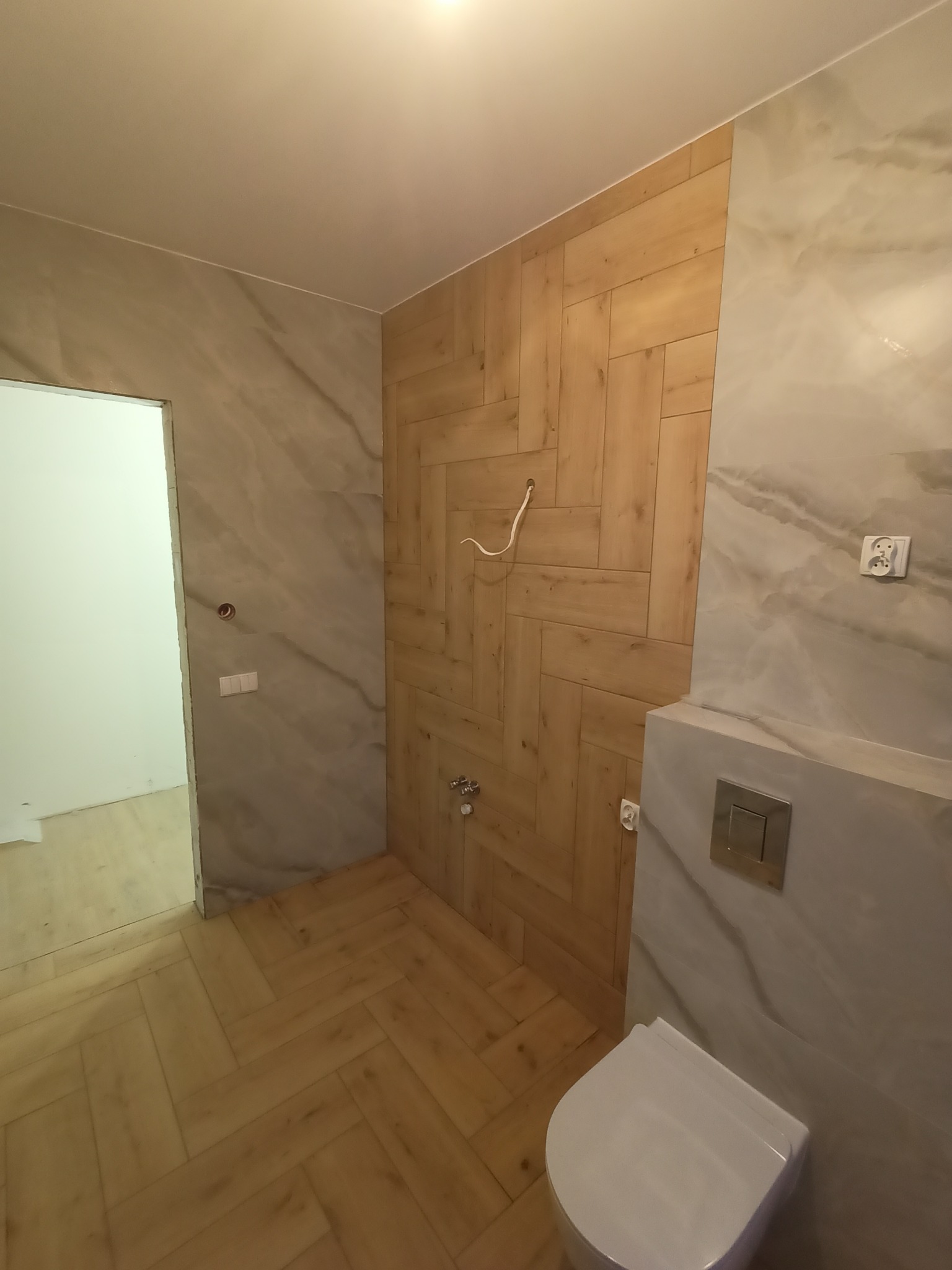 Nowoczesna łazienka z drewnopodobną okładziną ułożoną w jodełkę na ścianie i podłodze, marmurowymi płytkami na pozostałych ścianach, wiszącą toaletą i wystającym kablem elektrycznym.