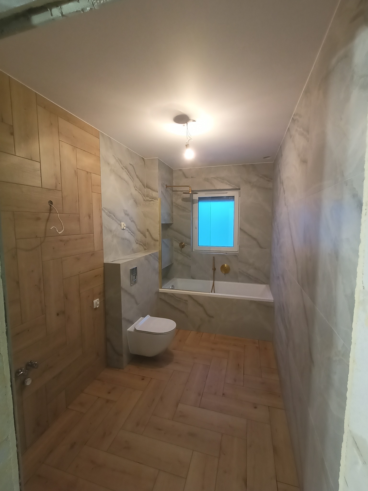 Wykończona łazienka z drewnopodobnymi płytkami ułożonymi w jodełkę na podłodze i ścianie, marmurowe płytki na ścianach, wisząca toaleta, wanna z niebieskim oknem i złotą armaturą.