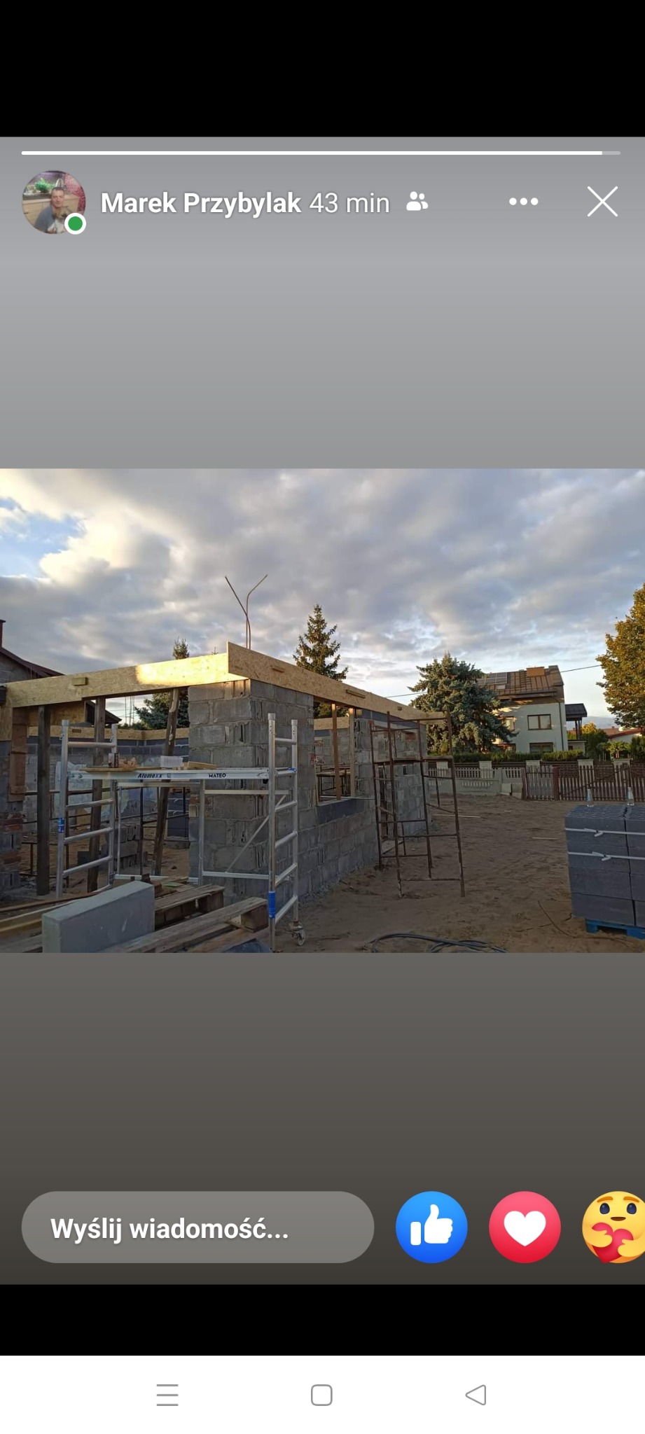 Budowa domu w trakcie realizacji: ściany z bloczków, drewniana konstrukcja dachu, rusztowania aluminiowe, widok na sąsiedni dom i drzewa w tle, pochmurne niebo.