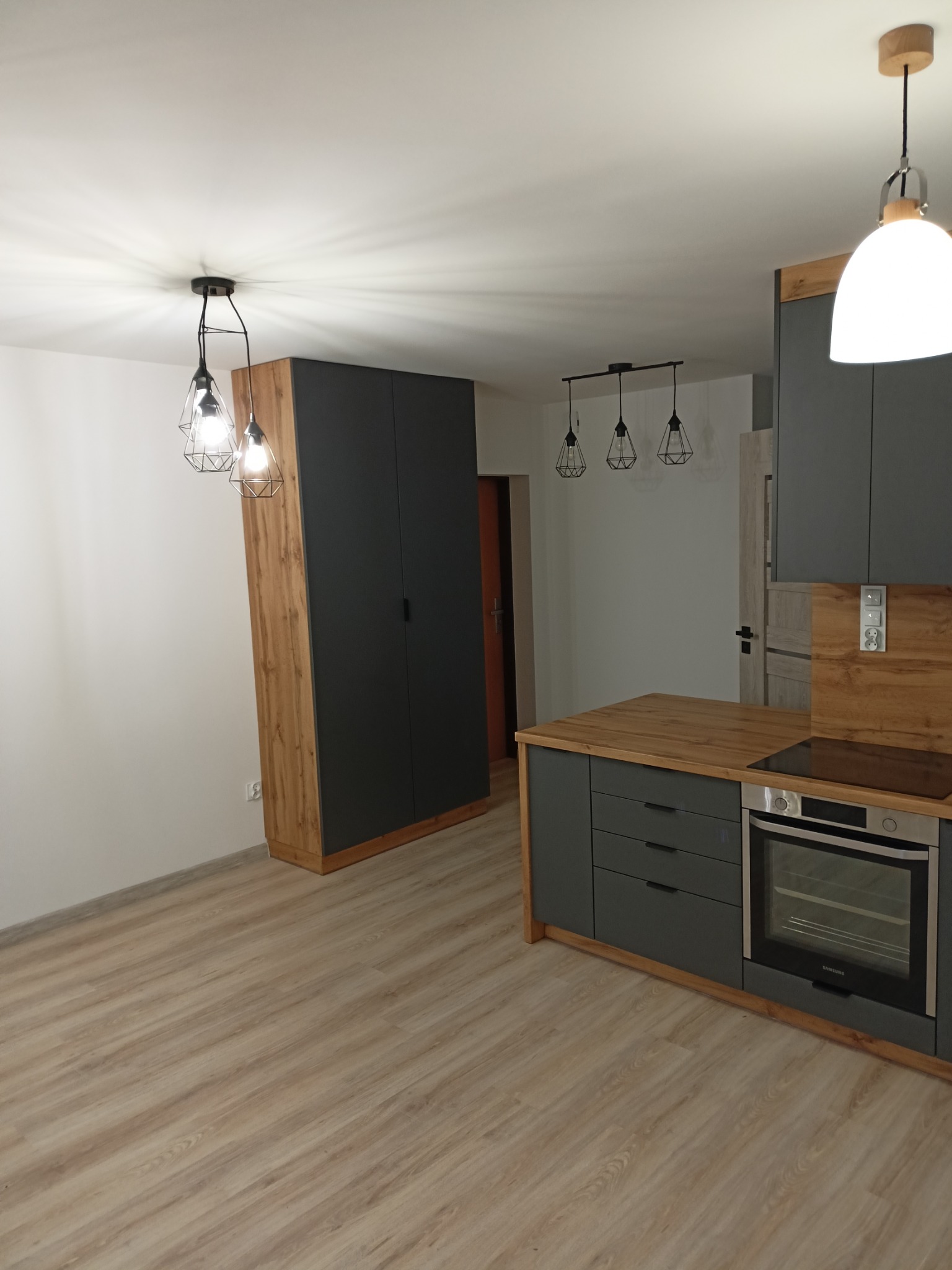 Nowoczesna kuchnia z szarymi szafkami, drewnianymi blatami i akcentami, oświetlona wiszącymi lampami o geometrycznych kloszach i pojedynczą lampą kulistą, z widoczną zabudową szafy w przedpokoju.