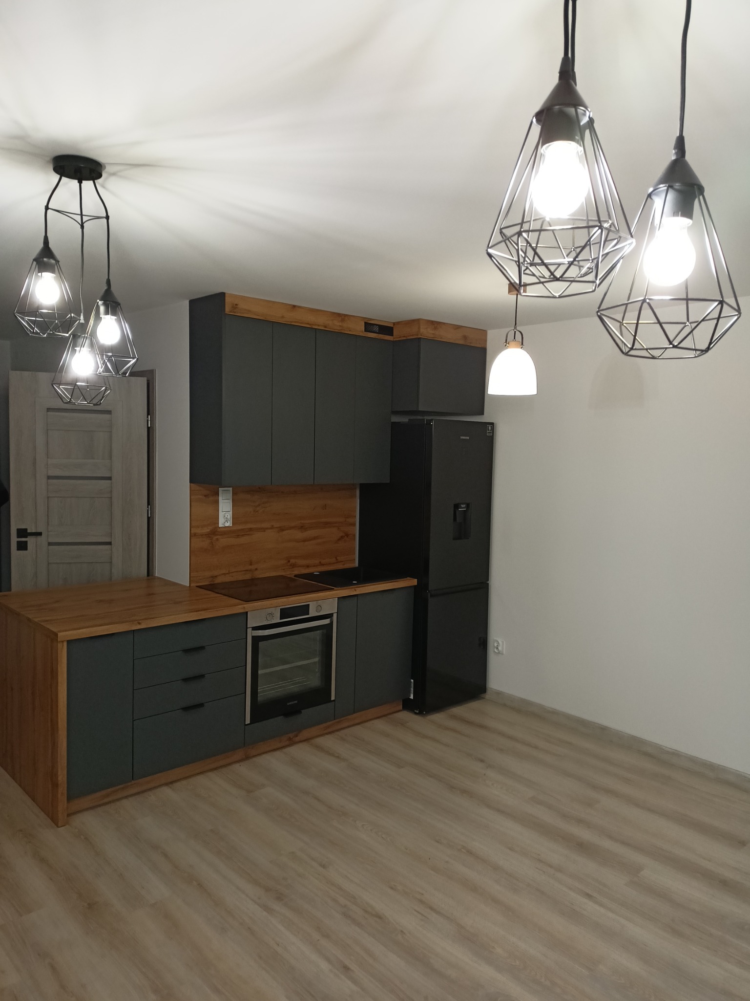 Nowoczesna kuchnia z szarymi szafkami, drewnianymi akcentami i designerskimi lampami wiszącymi w kształcie klatek, czarna lodówka w rogu, podłoga z paneli imitujących drewno.