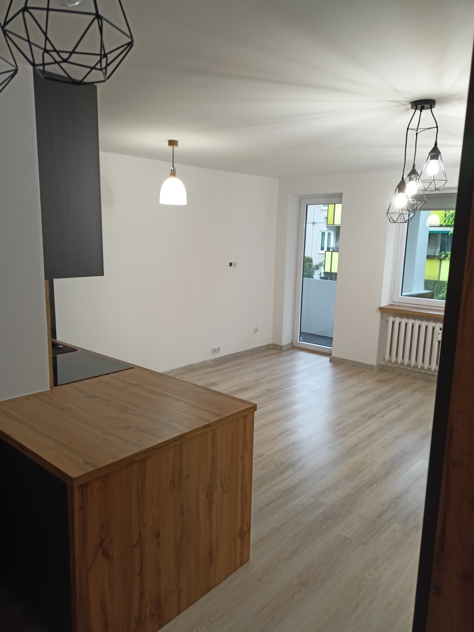 Jasne, przestronne wnętrze po remoncie z drewnianą podłogą, wyspą kuchenną z czarnym blatem, białymi ścianami i nowoczesnym oświetleniem.