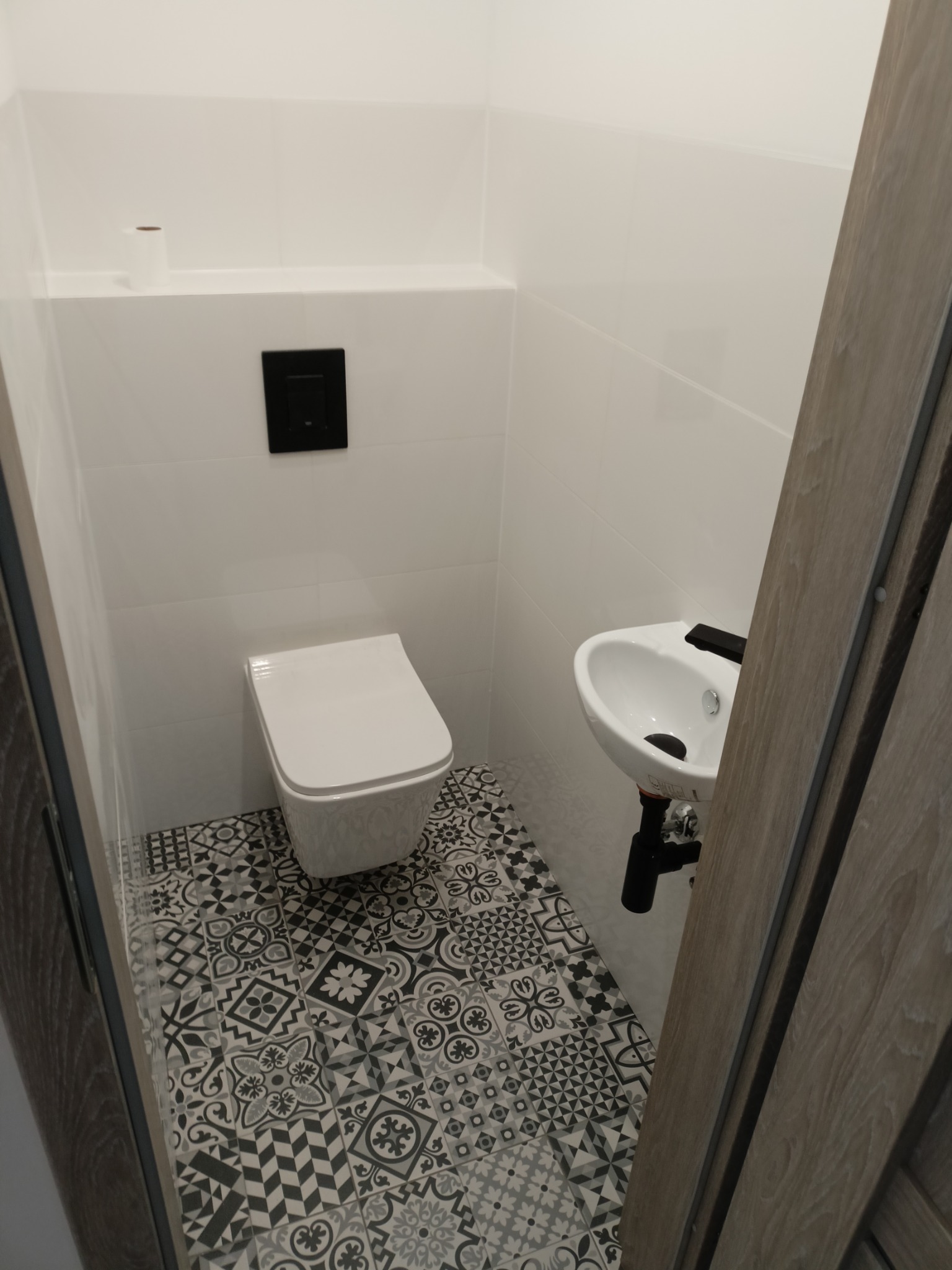 Mała łazienka z białą ceramiką, wzorzystą podłogą w odcieniach szarości i minimalistycznym czarnym przyciskiem spłuczki.