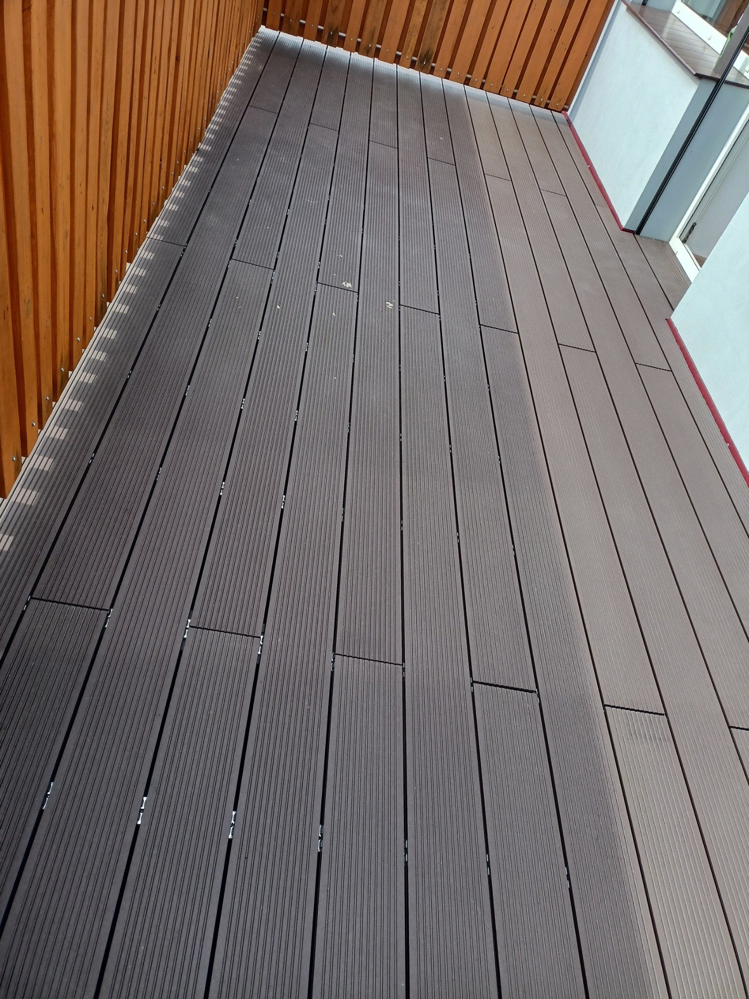 Balkon z szarych desek kompozytowych z widocznymi mocowaniami, obok drewniana ażurowa ścianka i biała ściana budynku z oknem.