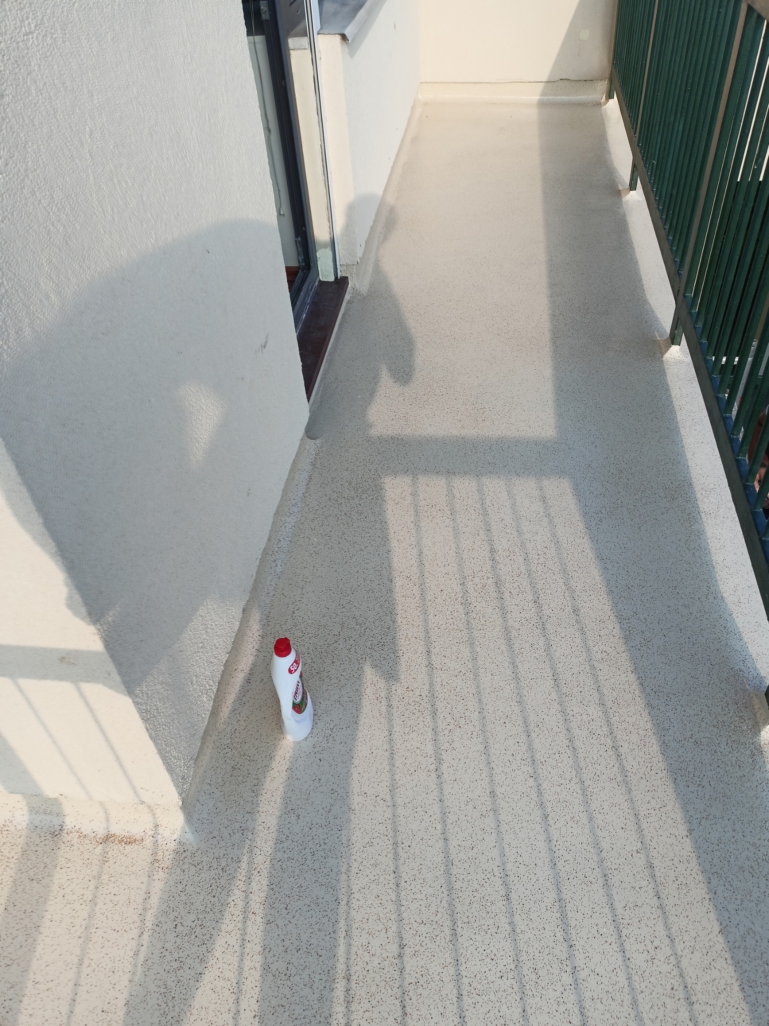 Biały balkon z szarą, nakrapianą posadzką, częściowo zacieniony przez balustradę, z butelką płynu do czyszczenia w rogu.