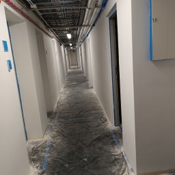 Mateusz Wiechowski - Długi korytarz w trakcie malowania, zabezpieczony folią i taśmą malarską, widoczne instalacje pod sufitem.