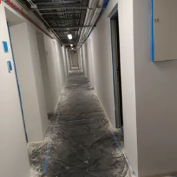 Długi korytarz w trakcie malowania, zabezpieczony folią i taśmą malarską, widoczne instalacje pod sufitem.