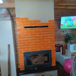 Mateusz Wiechowski - Malowanie kominka w cegiełki na biało i pomarańczowo, widoczne taśmy malarskie, wkład kominkowy z drewnem, puszka farby Dulux na podłodze, drewniane panele na ścianach.