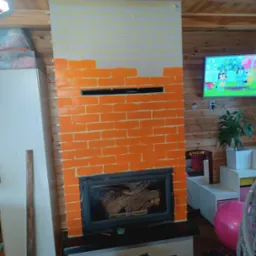 Malowanie kominka w cegiełki na biało i pomarańczowo, widoczne taśmy malarskie, wkład kominkowy z drewnem, puszka farby Dulux na podłodze, drewniane panele na ścianach.