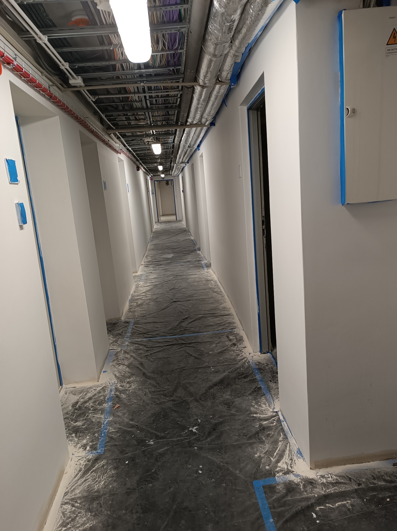 Długi korytarz w trakcie malowania, zabezpieczony folią i taśmą malarską, widoczne instalacje pod sufitem.
