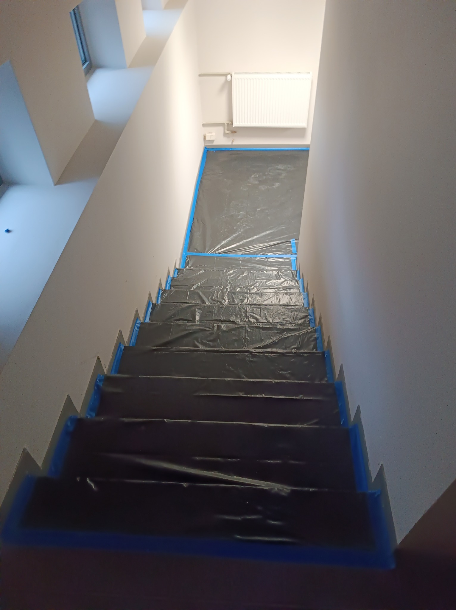 Schody zabezpieczone folią i taśmą malarską przed malowaniem ścian w jasnym wnętrzu, widok z góry.
