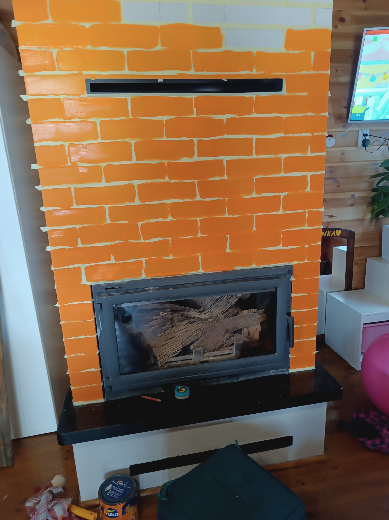 Malowanie kominka w cegiełki na pomarańczowo z użyciem taśmy malarskiej, widoczny wkład kominkowy z drewnem, puszka farby i narzędzia malarskie w pobliżu.