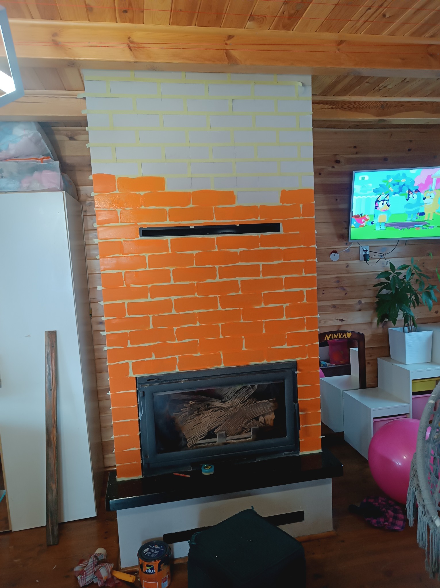 Malowanie kominka w cegiełki na biało i pomarańczowo, widoczne taśmy malarskie, wkład kominkowy z drewnem, puszka farby Dulux na podłodze, drewniane panele na ścianach.