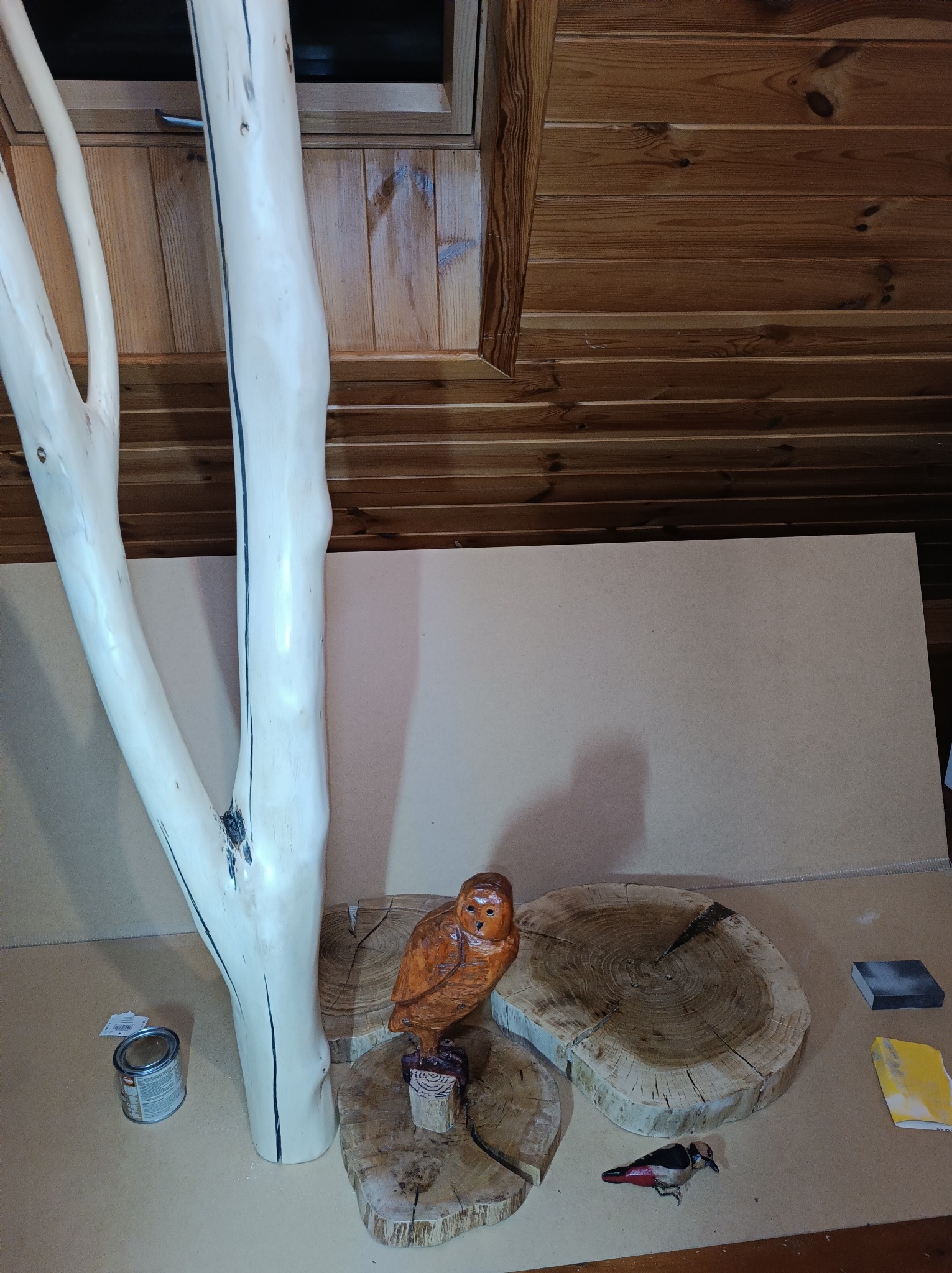 Ręcznie rzeźbiona drewniana sowa i dzięcioł na plastrach drewna, obok pobielonego pnia z czarnymi akcentami, puszka farby i kostki do szlifowania, na tle boazerii