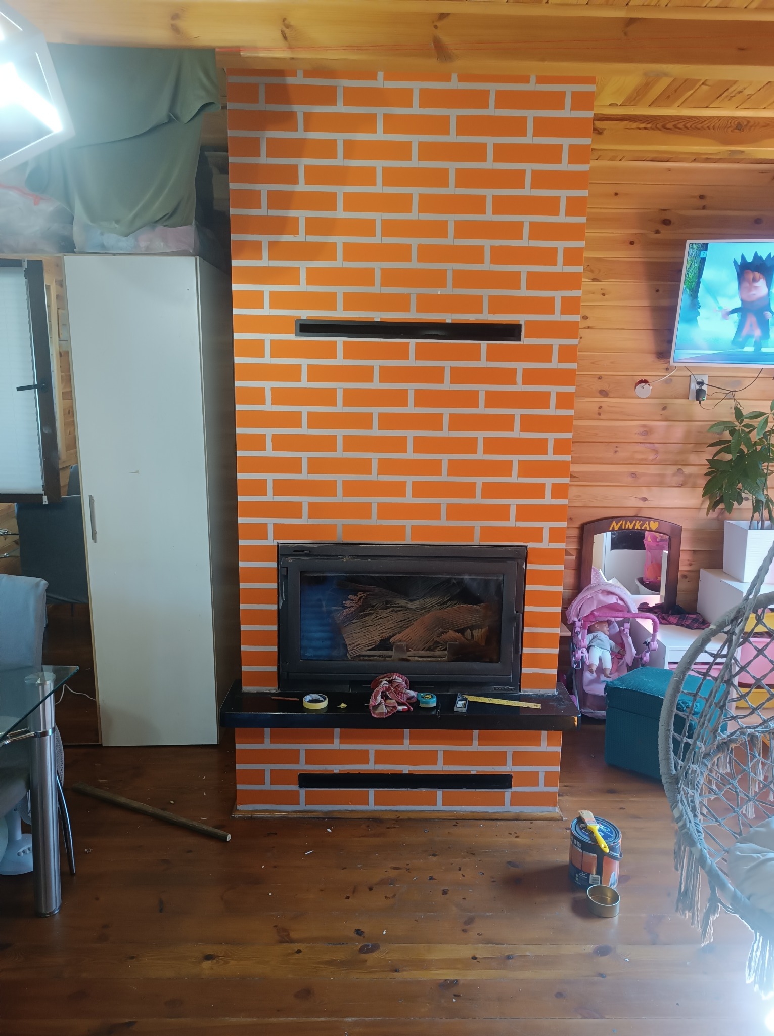 Pomarańczowy kominek z wzorem cegieł po malowaniu, widoczne narzędzia malarskie i resztki farby na podłodze w drewnianym domu.