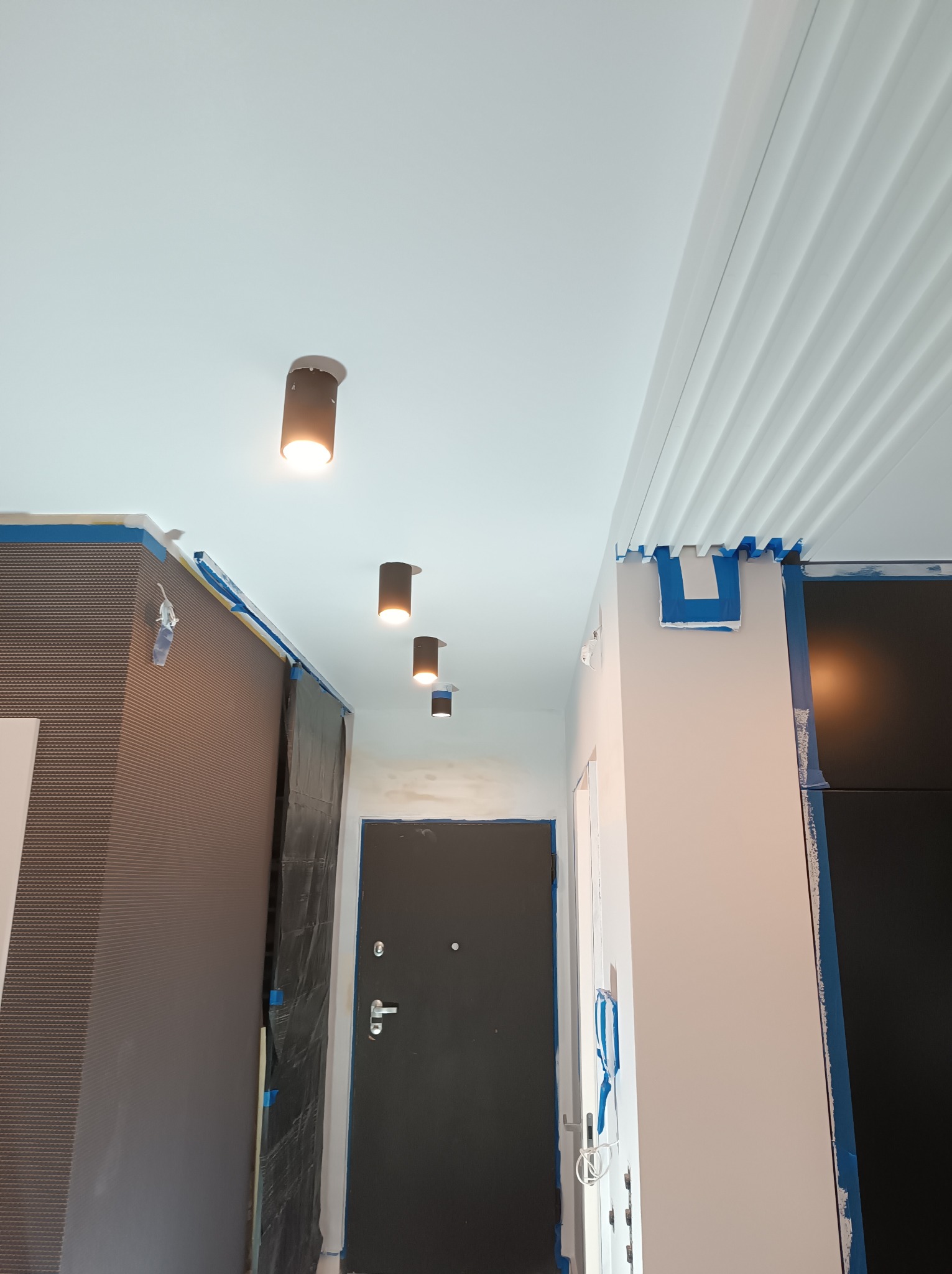 Korytarz w trakcie malowania: ściany zabezpieczone taśmą malarską, widoczne lampy sufitowe, drzwi wejściowe i fragmenty zabudowy meblowej.