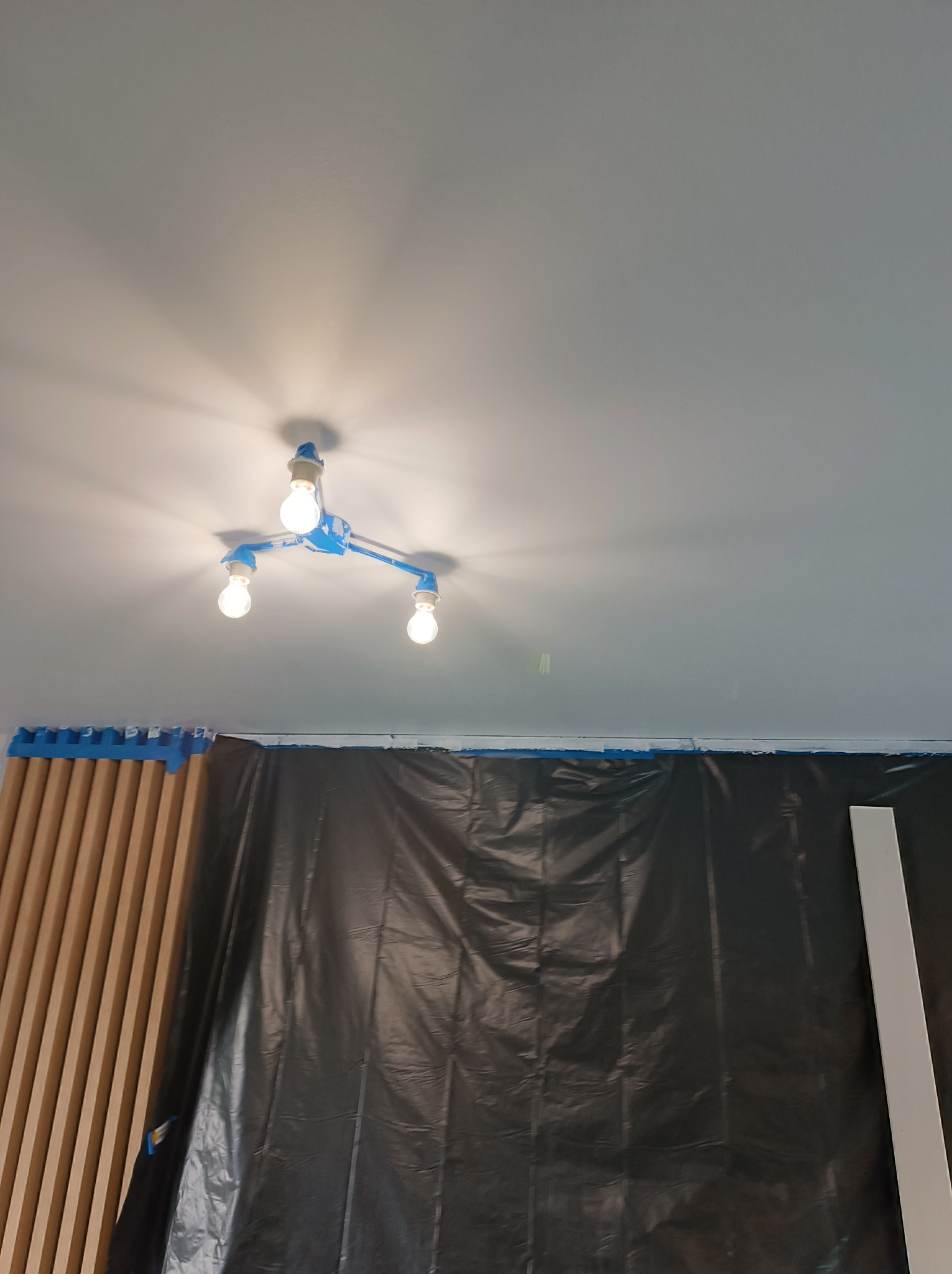 Przygotowania do malowania: sufit z lampą zabezpieczoną taśmą malarską, ściana osłonięta folią malarską, listwy przypodłogowe i panele ścienne zabezpieczone taśmą.