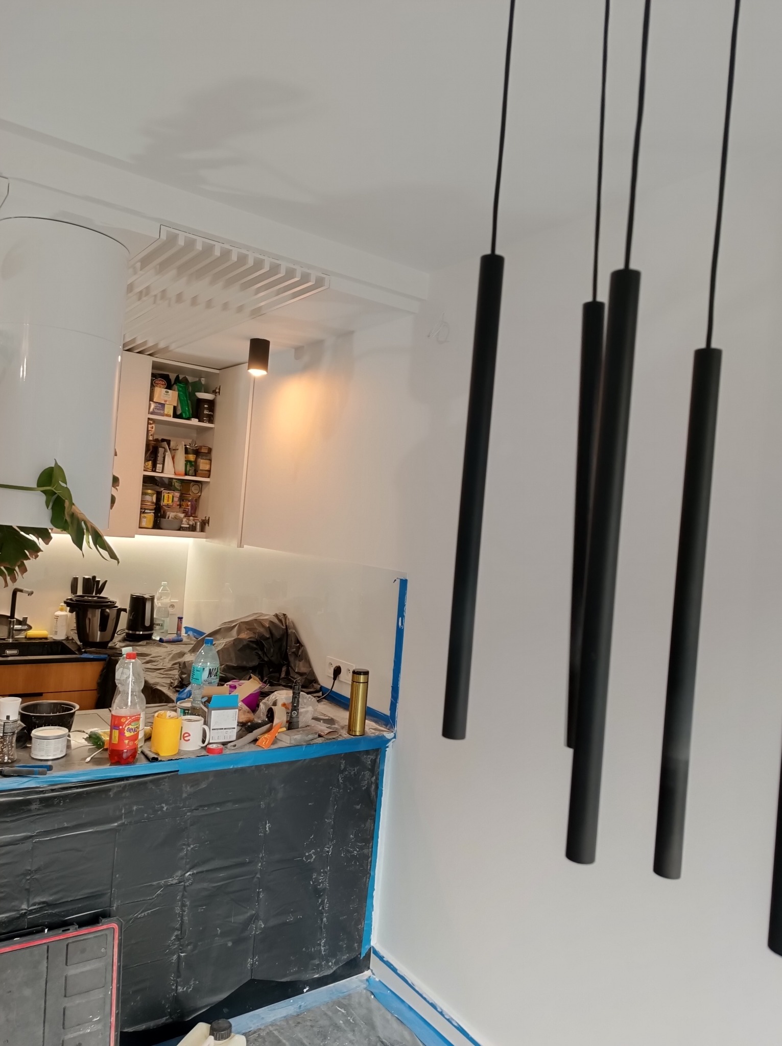 Wnętrze kuchni w trakcie prac wykończeniowych w Łochowie, zabezpieczone blaty, widoczne wiszące lampy rurowe i otwarte szafki.