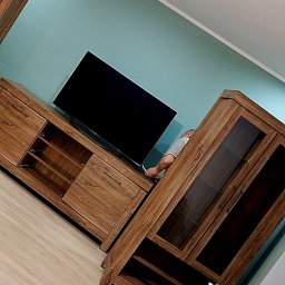 Budomex - Montaż mebli RTV z elementami szklanymi w salonie, widoczny pracownik podłączający telewizor do szafki RTV, na podłodze butelka wody i narzędzia