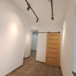 Jasne wnętrze z panelami podłogowymi w odcieniach brązu, przesuwnymi drewnianymi drzwiami i czarnymi reflektorami sufitowymi na szynoprzewodzie.