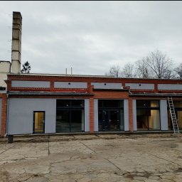 Budomex - Budynek po remoncie elewacji z szarą farbą, widoczna drabina oparta o ścianę, ceglane elementy dekoracyjne i wysoki komin w tle.