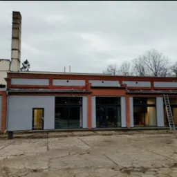 Budynek po remoncie elewacji z szarą farbą, widoczna drabina oparta o ścianę, ceglane elementy dekoracyjne i wysoki komin w tle.