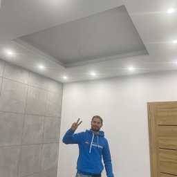 Budomex - Wnętrze pokoju po remoncie z podwieszanym sufitem z oświetleniem punktowym, szarą okładziną ścienną imitującą beton i drewnianymi drzwiami. Mężczyzna w niebieskiej bluzie z logo Atlas pozuje...