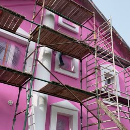 Budomex - Różowa elewacja domu w trakcie odnawiania; widoczne rusztowania, okna zabezpieczone folią, pracownik przy oknie.