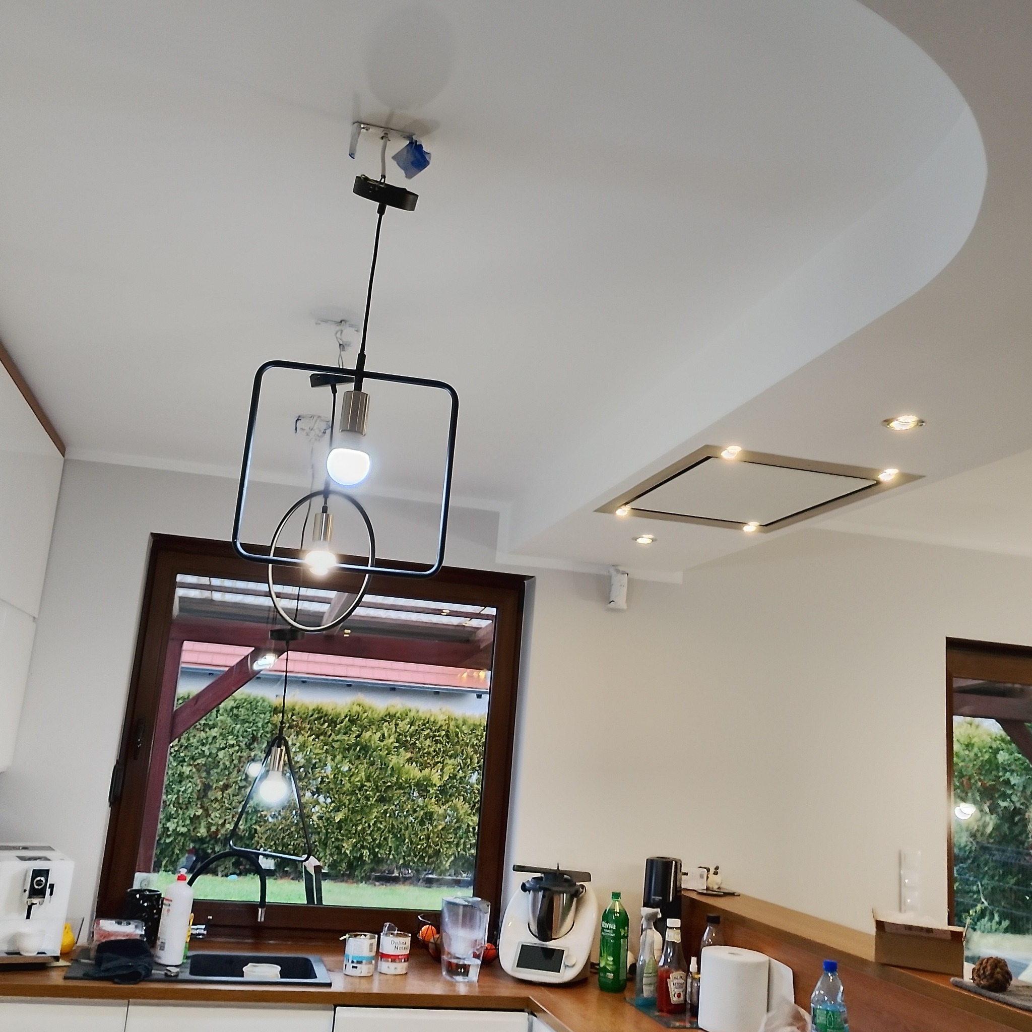 Nowoczesna kuchnia z oknem na ogród, wisząca lampa z geometrycznym kloszem i podwieszany sufit z oświetleniem punktowym.