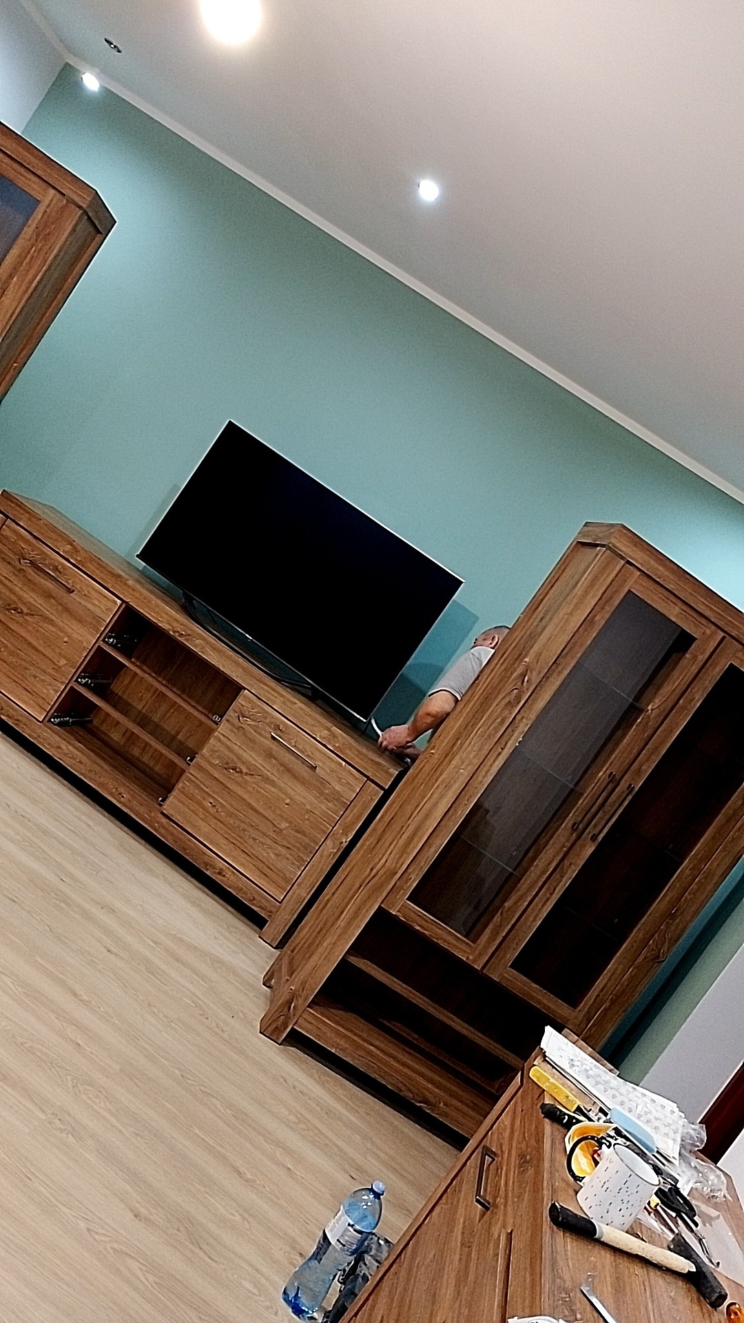 Montaż mebli RTV z elementami szklanymi w salonie, widoczny pracownik podłączający telewizor do szafki RTV, na podłodze butelka wody i narzędzia