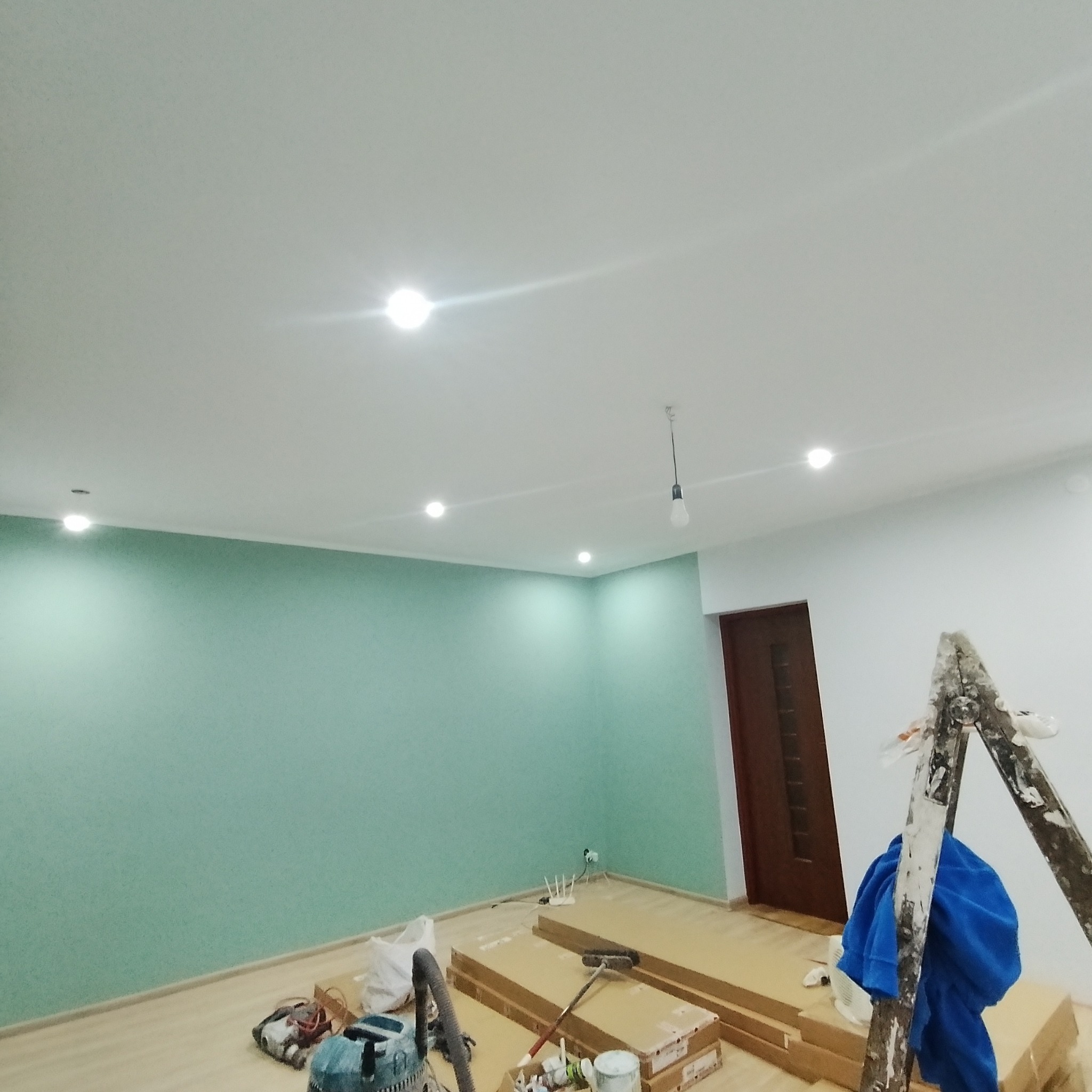 Pokój w trakcie prac wykończeniowych: ściana pomalowana na odcień zieleni, widoczna drabina malarska, sprzęt do szlifowania, listwy przypodłogowe w kartonach oraz wisząca żarówka na tle białego...